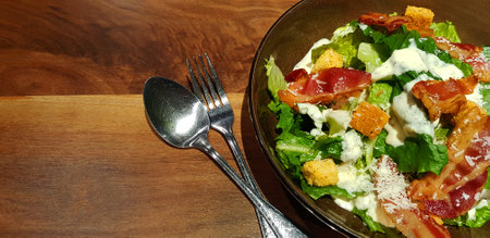 Fresh Caesar salad with vegetable, fried bacon, crispy bread, spoon and fork on brown wooden background or table with copy space. Healthy food topping with cream sauce. Good lifestの写真素材