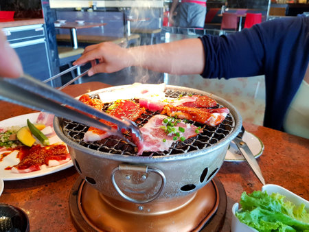Asian woman grilled raw sliced beef and pork on the hot stainless steel stove with smoke. Barbecue or Lunch time and Japanese style yakiniku. Selective focus on food.の写真素材