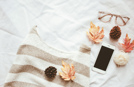 Autumn fashion style concept, sweater and smartphone with maple leaves on white bed sheet backgroundの写真素材