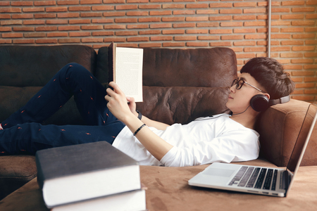 Young asian college man lying on a couch, reading a textbook and listening music from headphones in modern living roomの写真素材