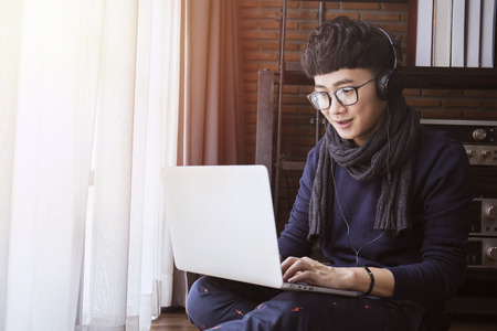 Young asian student wearing sweater studying on computer laptop and listening to music in living...