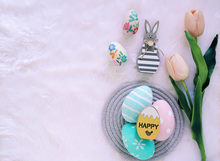 Happy Easter concept with wooden bunny and colorful easter eggs on white fur background and pink tulips. Top view with copy spaceの写真素材