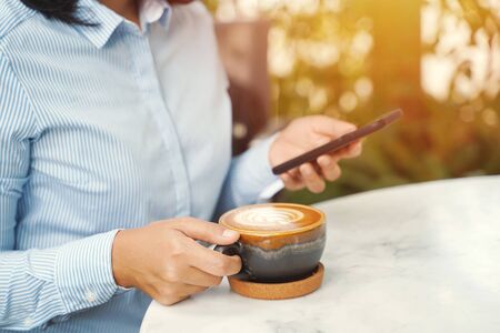 Close up of businesswoman using smartphone and drinking hot latte coffee in the morning at the cafe, business and lifestyle conceptの写真素材