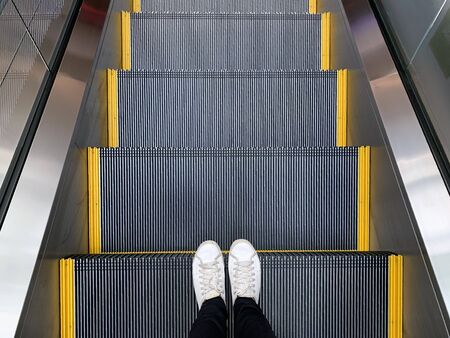 Selfie of feet in white sneakers shoes standing on escalator in shopping mall or modern officeの写真素材