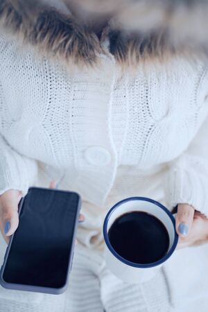 Top view and close up woman hands holding hot cup of coffee and smartphone in cold weather background, wearing warm fur knitted clothesの写真素材