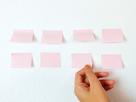 Woman hand picking blank pink sticky note on white wall background in office, business and presentation idea の写真素材