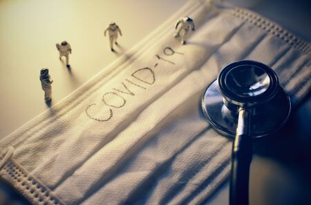 Miniature people doctors with protective suit prevention of pandemic Covid-19 and Coronavirus, surgical mask with Covid-19 text written on it with stethoscope, dramatic and dark tonedの写真素材
