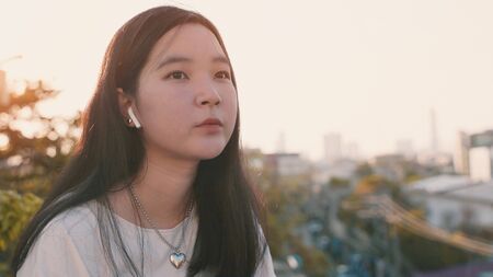 Young asian millennial gen z woman standing at rooftop feeling bored and looking at cityscape while listening to music, self quarantine or stay at home during COVID-19 and coronavirus pandemicの写真素材