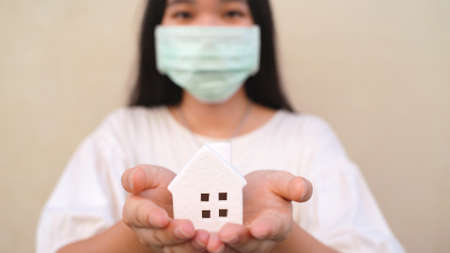 Young asian millennial gen z woman holding white house model and wearing mask for self quarantine or stay at home concept during COVID-19 and coronavirus pandemicの写真素材