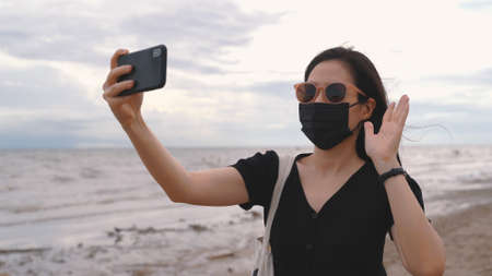 Young asian woman wearing medical mask and video call   by smartphone at the beach after coronavirus, new normal lifestyle and travel bubble conceptの写真素材