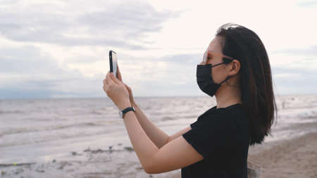 Young asian woman wearing medical mask and taking photos by smartphone at the beach after coronavirus, new normal lifestyle and travel bubble conceptの写真素材