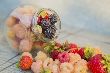 Black, red, yellow raspberries in a glass jar with strawberries on a old wooden backgroundの写真素材