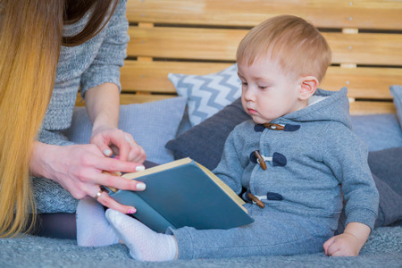 Young mother and her baby son sitting on the bed and reading book togerher at home.の写真素材