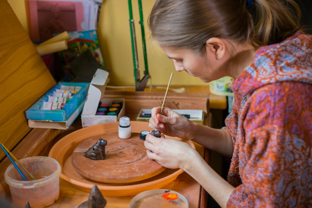 Professional woman potter, decorator painting ceramic souvenir penny whistle toy dog in pottery...
