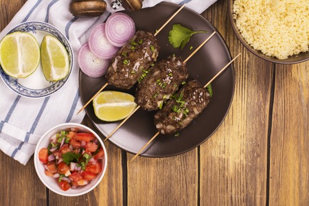 Grilled kebabs with vegetable salad, couscous and seasonings over wooden table