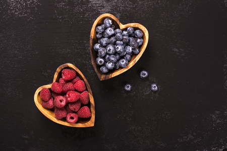 Blueberry and raspberry in heart shaped wooden bowls over black stone background. Top viewの写真素材