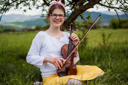 Beautiful blonde girl sitting on a beautiful green grass full of flowers and enjoy the sounds of the violin, girl playing the violin in the countryside on a beautiful meadowの写真素材