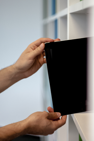 Guy or man or student or businessman taking a tablet with a blank screen from a bookshelf in the library with his handの写真素材