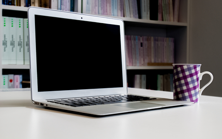 Laptop with a blank screen and colorful coffee mug on the desk with bookshelves in the background in...