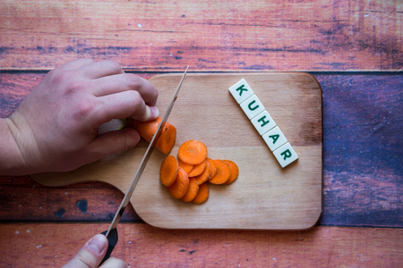 Chef is cutting carrot on a wooden cutting board with sharp knife and word for bosnian, serbian, croatian chef written with box letters.の写真素材