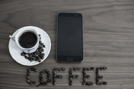 Roasted coffee beans, scattered on the table and written Coffee, in a cup of roasted, over coffee, and a mobile phone, brown wood texture, background wood, plates of tableの写真素材