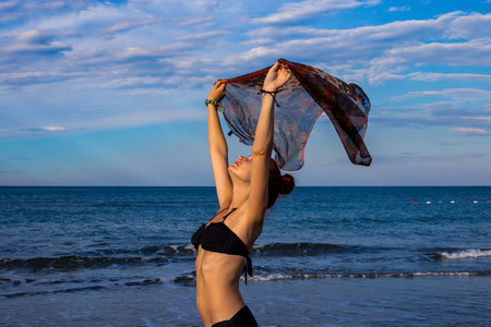 Slim good looking tanned redhead with arms outstretched holding scarf while wind is blowing on the beach in black bikini with blue sea and sky in the background during summerの写真素材
