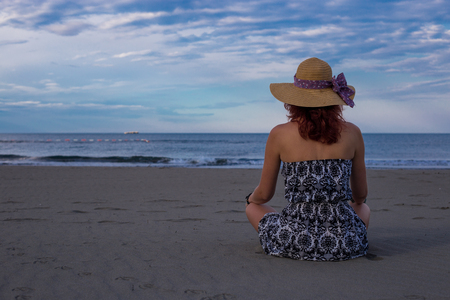 Beautiful girl woman in dress and hat sitting on the beach with back turned and sea in the backgroundの写真素材