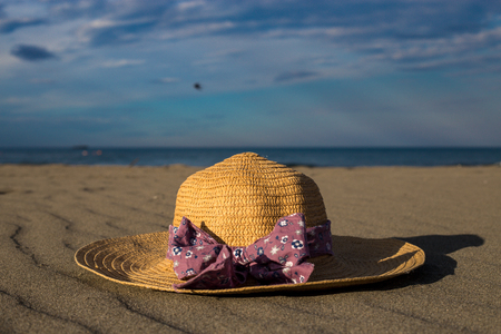 Beautiful hat closeup with purple bow ribbon laying on a dreamy sand beach with blue sky and sea in the backgroundの写真素材
