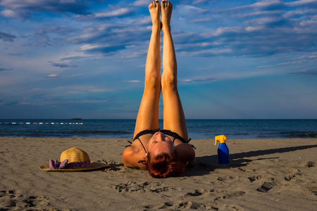 Young good looking tanned redhead laying on a sandy beach with arms resting and legs raised in the air posing and resting with sun cream and hat on the side and blue sky and sea in the background during summer の写真素材