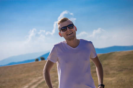 Handsome young caucasian blonde guy happy man with reflective black sunglasses laughing and smiling. Grass field in the summer with mountains and blue sky and clouds in the backgroundの写真素材