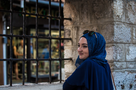 Young and beautiful caucasian covered muslim girl with blue hijab and sunglasses looking through black metal bars near mosque with old stone wall and people walking in the backgroundの写真素材