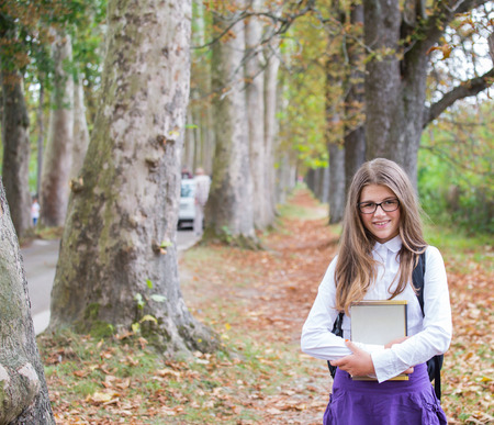 Pretty beautiful blonde child schoolgirl back to school standing and smiling in tree alley in autumn nature holding notes and books with backpack, glasses, white shirt and purple skirtの写真素材