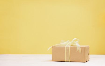 Gift box wrapped in craft paper and tied with yellow ribbon on white wooden table and yellow...
