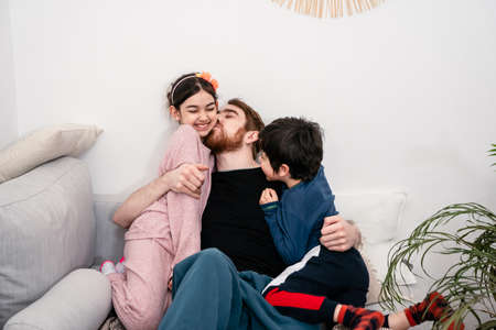 Father and two kids embracing and having fun on sofa at home. Multinational family. Real life, togetherness, diversity, home comfortの写真素材