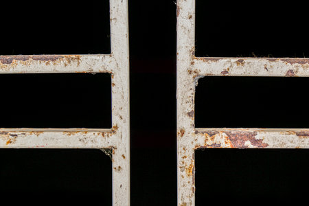 Close-up of a weathered white metal gate with rust, creating a textured pattern against a dark...