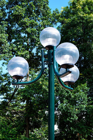 A green lamppost with four globe lights stands tall against a backdrop of lush green trees and a...