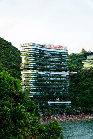 Flamingo resort, a modern green building with vertical gardens, overlooks a crowded beach where tourists enjoy swimming and sunbathing on a sunny day in a tropical coastal destination.のeditorial素材