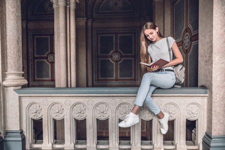 Beautiful and confident. Young female student sitting on the railing in the university hall and reading book.の写真素材