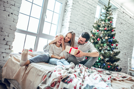 Christmas. Family. Happiness. Dad is giving a present to his daughter who is sitting on her mom's knees. At home near the Christmas tree, all are smilingの写真素材