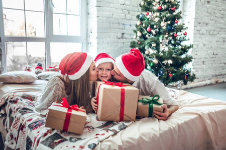Happy parents kissing daughter and giving xmas present on bed. Christmas tree backgroundの写真素材