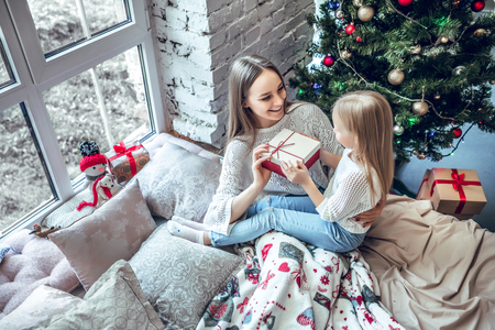 Cheerful mom and her cute daughter girl exchanging gifts near window. Parent and little child having fun near Christmas tree indoors. Morning Xmas. top viewの写真素材