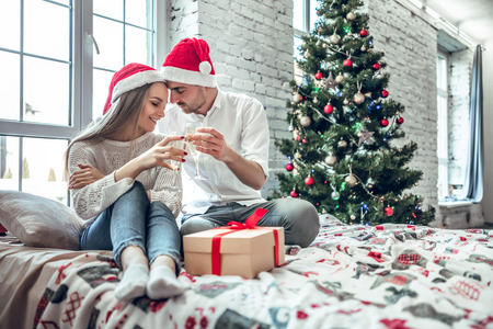 Beautiful couple in love, wearing santa hats, sitting next to a nicely decorated Christmas tree and making a toast with glasses of champagne.の写真素材
