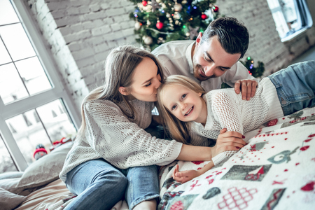 A family of three hugging in front of the christmas tree on the bed. New year's eve. Cozy holiday at the fir-tree. love, happiness and big family conceptの写真素材