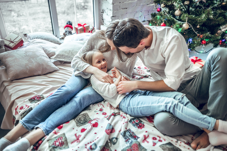 Merry parents and girl child together make plans for Christmas holidays and lie on large bed in beautiful bright bedroom with Christmas tree.の写真素材