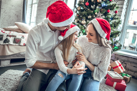 People, family, celebration and holidays concept. Mother, father and daughter with attractive appearance look at each other s eyes, sit on white carpet near decorated New Year tree and gift boxの写真素材