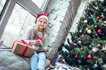 Cute little baby girl sitting by the window. Room decorated on Christmas. Kid enjoys the giftの写真素材