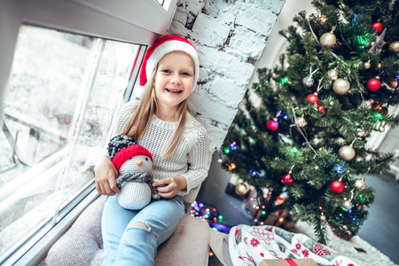 Cute little girl sitting by the window and looking at the camera. Room decorated on Christmas.の写真素材