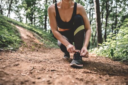 Cropped image of young athlete woman preparing to running outdoor on the forest path. Slim athletic girl ties up shoelaces on shoes before training in the park.の写真素材