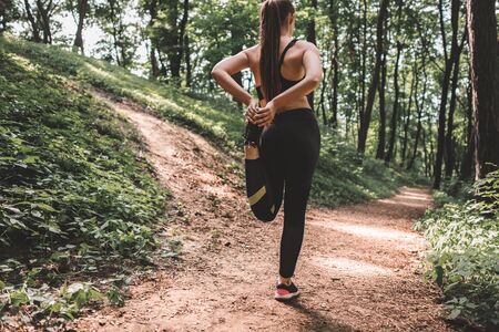 Full length image of young woman athlete making warm-up before training in the woods. Back view of a slim fit female preparing to run on forest pathの写真素材