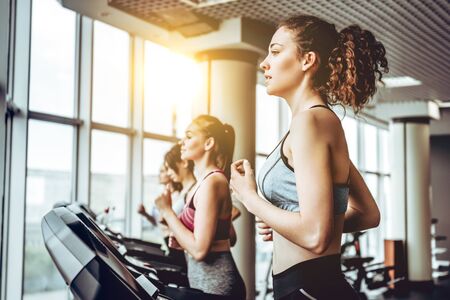 Sporty beauty. Beautiful young cheerful woman in sportswear running on treadmill at gym with other women.の写真素材